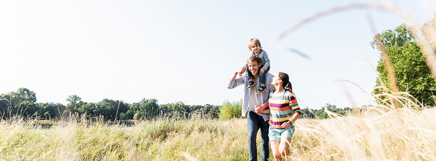 Husband and wife holding hands while walking through a field with a child sitting atop the father's head shoulder